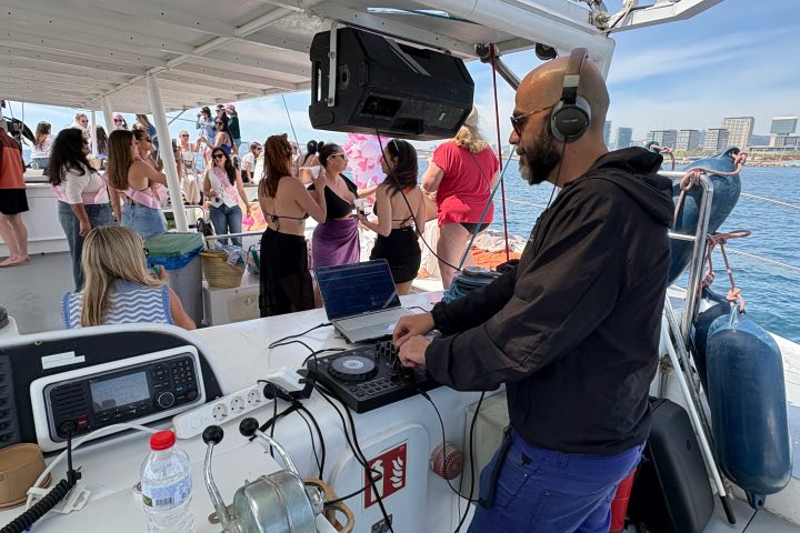 DJ with headphones performing on a boat, crowd in the background enjoying the music.